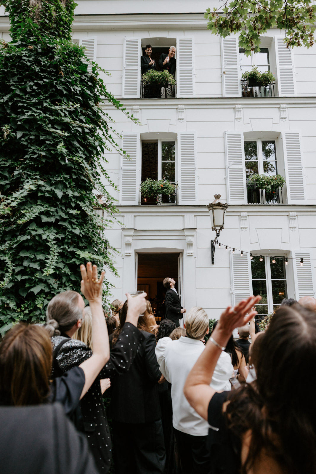 the grooms pop their heads out of one of the suites, saying hi to their guests below just before the ceremony starts at this gay wedding in the garden of Hotel Particulier Montmartre, Paris